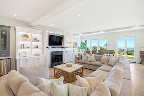 A spacious living room with a large sectional sofa, a flat-screen TV mounted above a fireplace, and a view of the ocean through large windows. - Port of Call on Isle of Palms OCEAN FRONT, Pool, boardwalk, putting green (Isle of Palms)