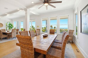 A dining area with a wooden table and wicker chairs, large windows with a view of the ocean, and a ceiling fan. - Port of Call on Isle of Palms OCEAN FRONT, Pool, boardwalk, putting green (Isle of Palms)