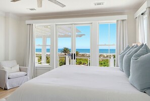 Un dormitorio con una cama grande, una silla y vista al mar a través de ventanas abiertas.