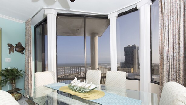 Ein Balkon mit einem Glastisch, weißen Stühlen und Blick auf Stadt und Meer.