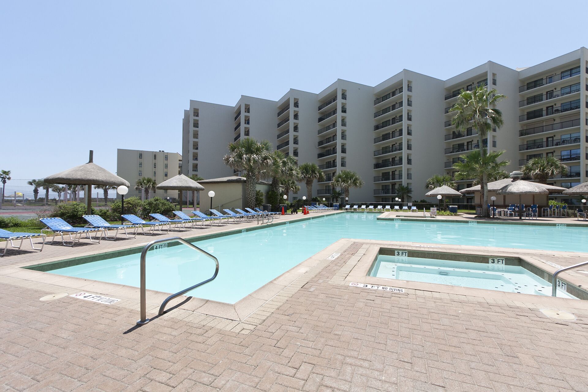 A swimming pool with lounge chairs and umbrellas, surrounded by a multi-story building with balconies.