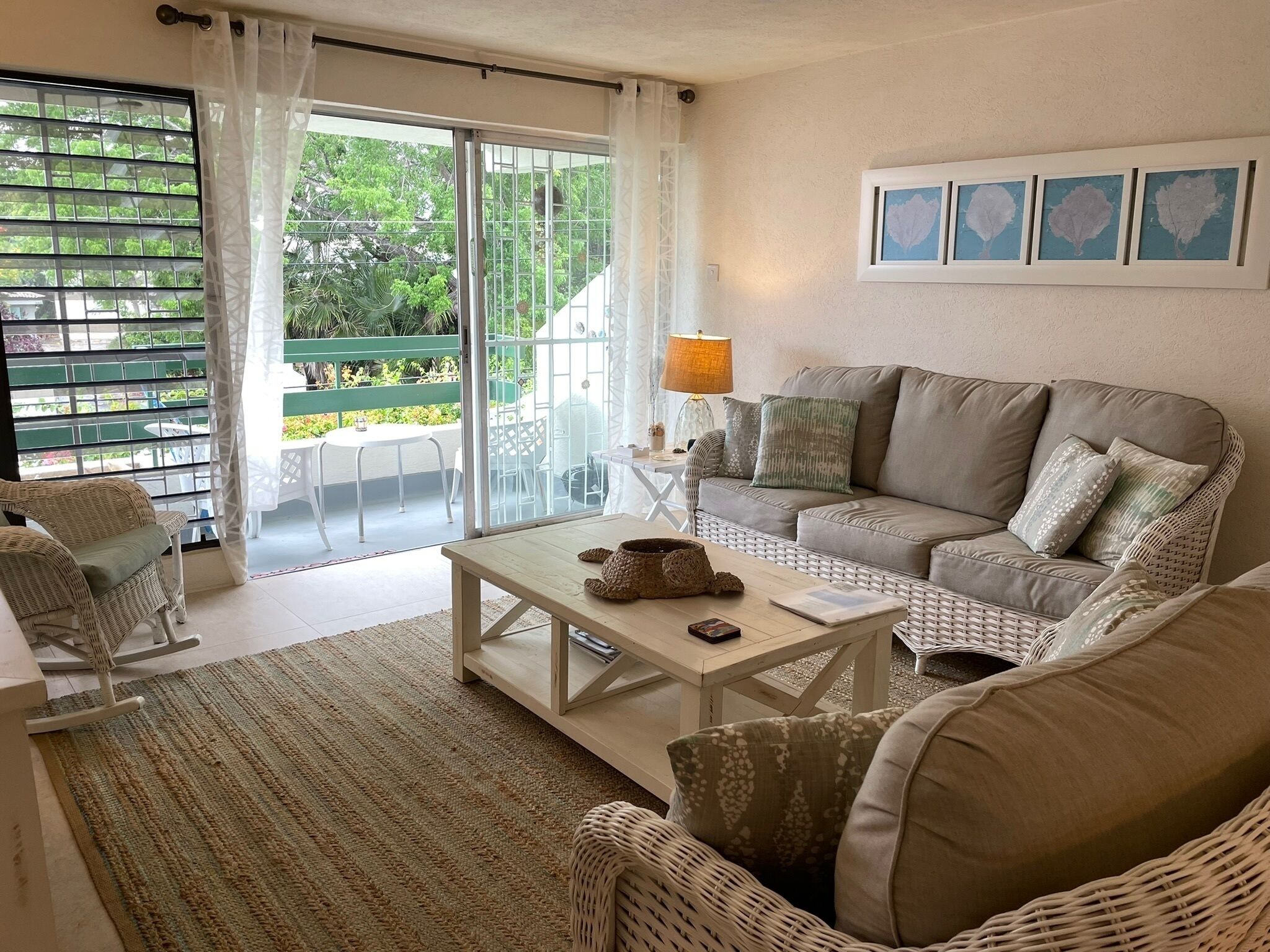 Un salon chaleureux avec un canapé, une table basse et un fauteuil en osier. Il y a un balcon avec vue sur la verdure.
