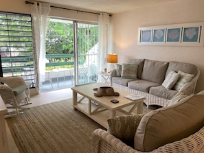 A cozy living room with a sofa, a coffee table, and a wicker chair. There is a balcony with a view of greenery. - Banyan Court, a Family-Friendly Condo Close to Everything! (Barbados)