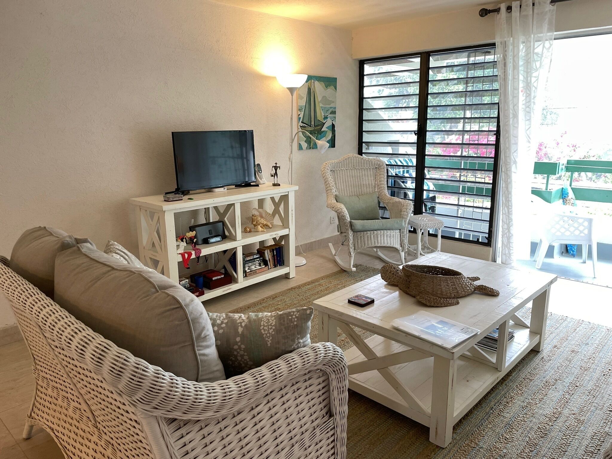 A living room with a white wicker sofa, a white coffee table, a television on a white cabinet, a rocking chair, and a view of a balcony with a railing.