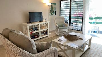 A living room with a white wicker sofa, a white coffee table, a television on a white cabinet, a rocking chair, and a view of a balcony with a railing.