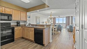 Una cucina moderna con mobili in legno, forno integrato e lavastoviglie. C'è una zona pranzo con tavolo e sedie, e vista sull'oceano attraverso ampie finestre.