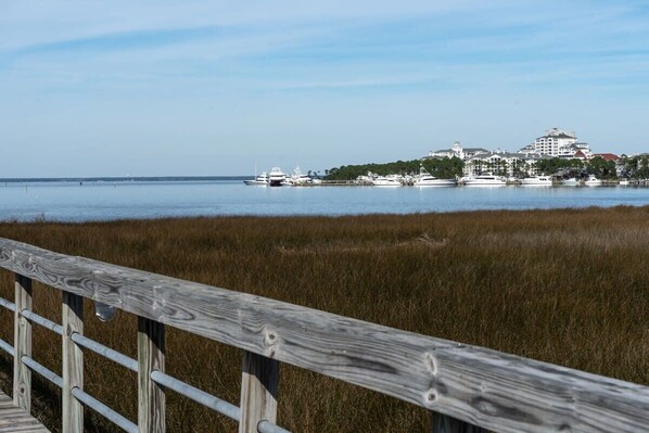 A coastal view with a wooden boardwalk leading to a marina with yachts and buildings in the background.