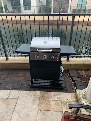 A portable grill on a patio with a railing and a table in the background.