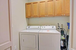 A laundry room with two stacked front-loading washers and dryers, wooden cabinets above, and a blue cleaning cart. - Luxury at 2,000+ SqFt! Baytowne and Pool! Golf Cart! Pond View! - Poppy's Place at Villa Lago Sandestin® (Miramar Beach)