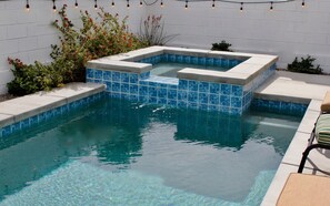 Un jacuzzi rectangular con mosaicos azules, rodeado por un deck de concreto y una pared de ladrillo blanco.