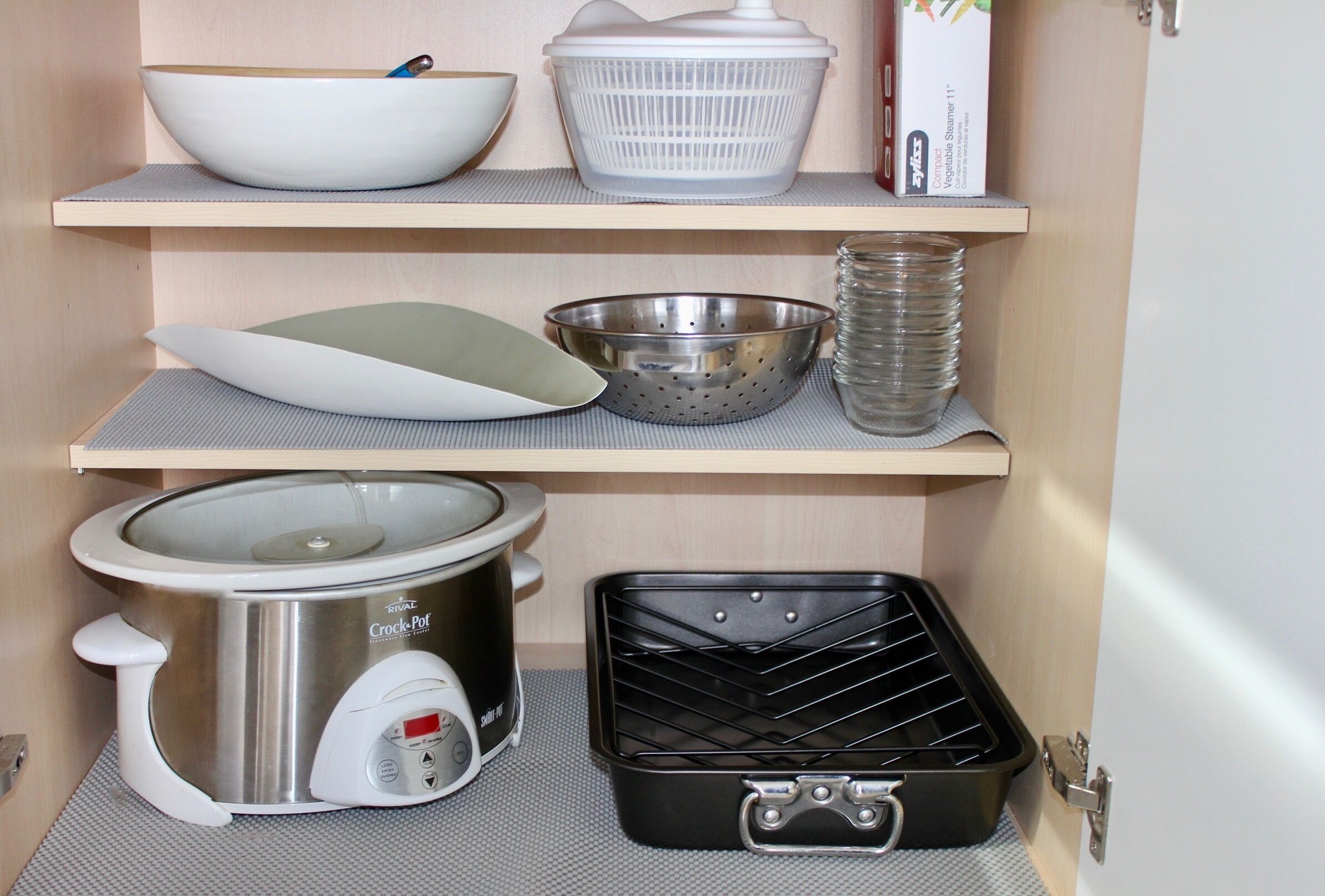 A kitchen with a slow cooker, colander, bowls, and a stack of clear glasses on shelves.