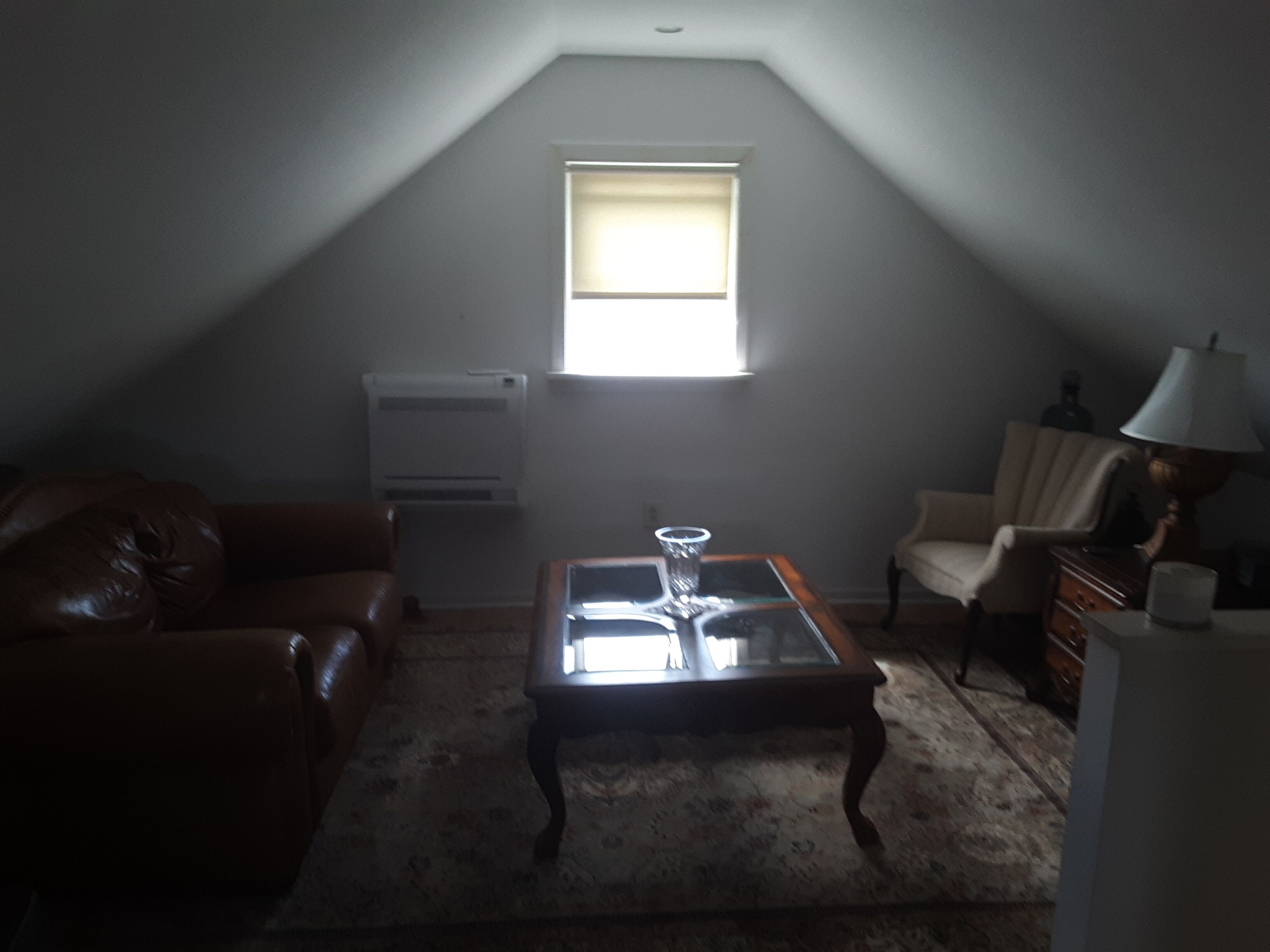 A dimly lit attic room with a sloped ceiling, a single window, a leather couch, a glass-top coffee table, a chair, and a lamp.
