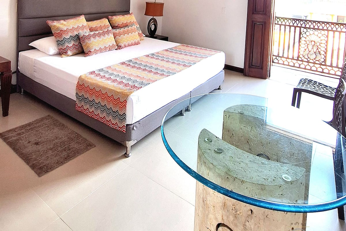 A bedroom with a bed, a nightstand, a lamp, a glass table, and a balcony with a railing.