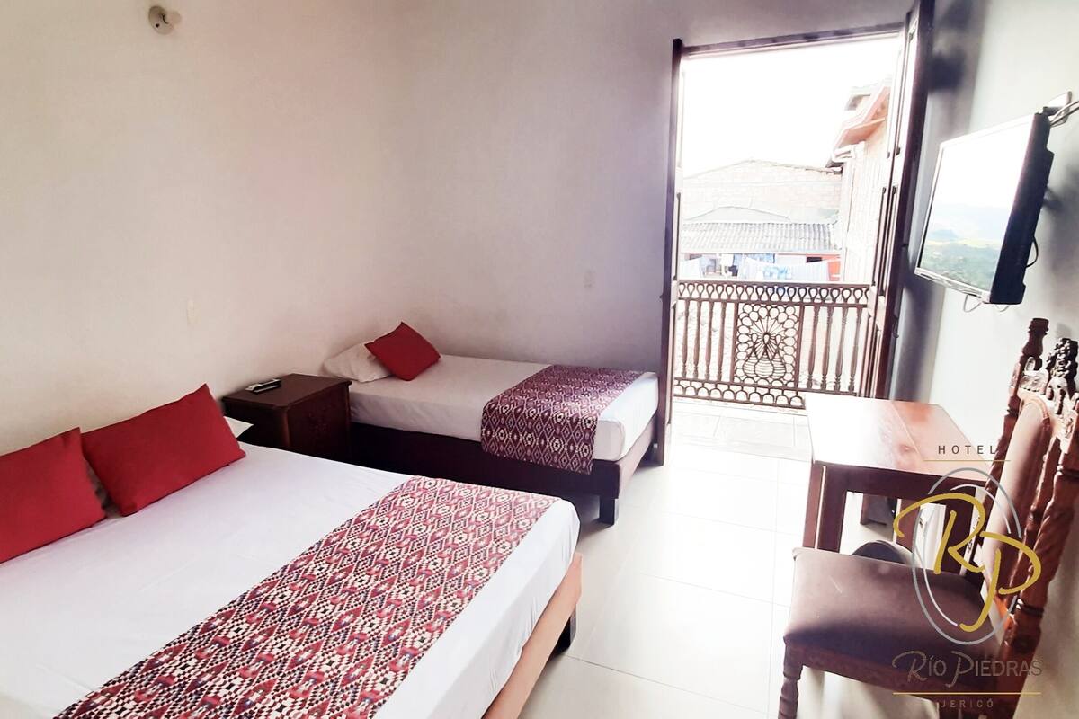A hotel room with two beds, each with red pillows and patterned bedspreads. There is a wooden nightstand with a remote control, a small table with a chair, and a TV mounted on the wall.