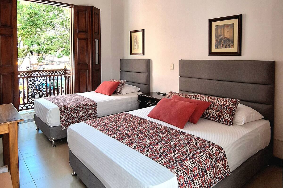 A hotel room with two beds, a balcony, and a view of greenery outside.