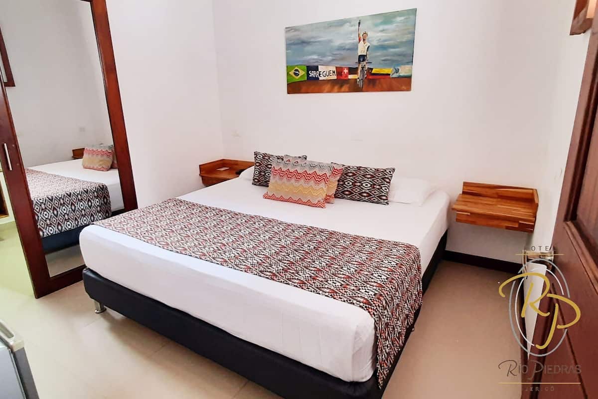 A double bed with patterned bedding, a bedside table, a nightstand, a mirror, a bathroom with a sink and mirror, and a painting on the wall.