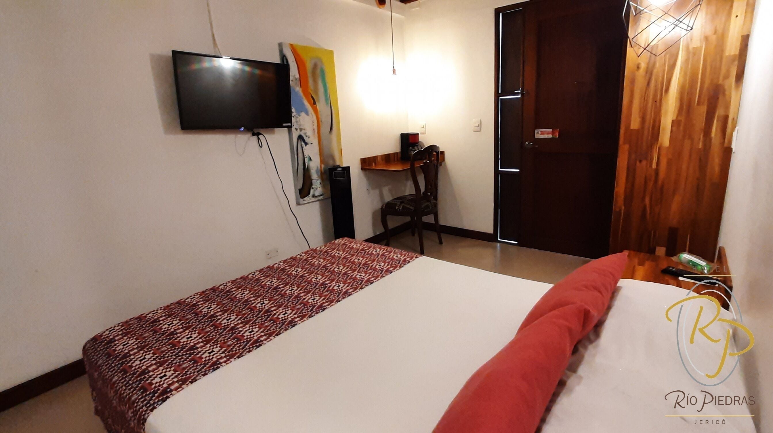Ett hotellrum med en säng, en tv som är monterad på väggen, ett skrivbord med en stol och en dörr.