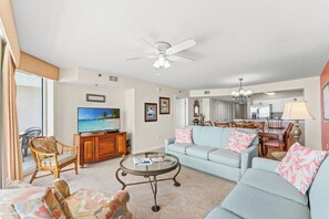A living room with a television, a ceiling fan, a dining area, and a view of the ocean.