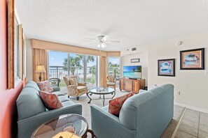A living room with a sofa, armchairs, a coffee table, and a TV. There is a balcony with a view of palm trees and the ocean.