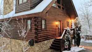 A log cabin with a stone foundation, a chimney, and a snow-covered roof.