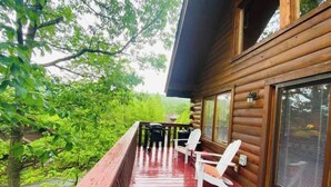 A wooden cabin with a red deck, white Adirondack chairs, and a large window overlooking greenery.