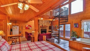A cozy cabin interior with a bed, a dining area, a fireplace, a spiral staircase, and a view of the outside.