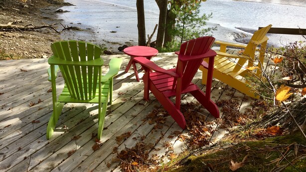 Terraza de madera con sillas Adirondack y una pequeña mesa, con vistas a un lago rodeado de follaje otoñal.