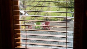 Una ventana con persianas horizontales, vistas a un paisaje verde y un patio con sillas.