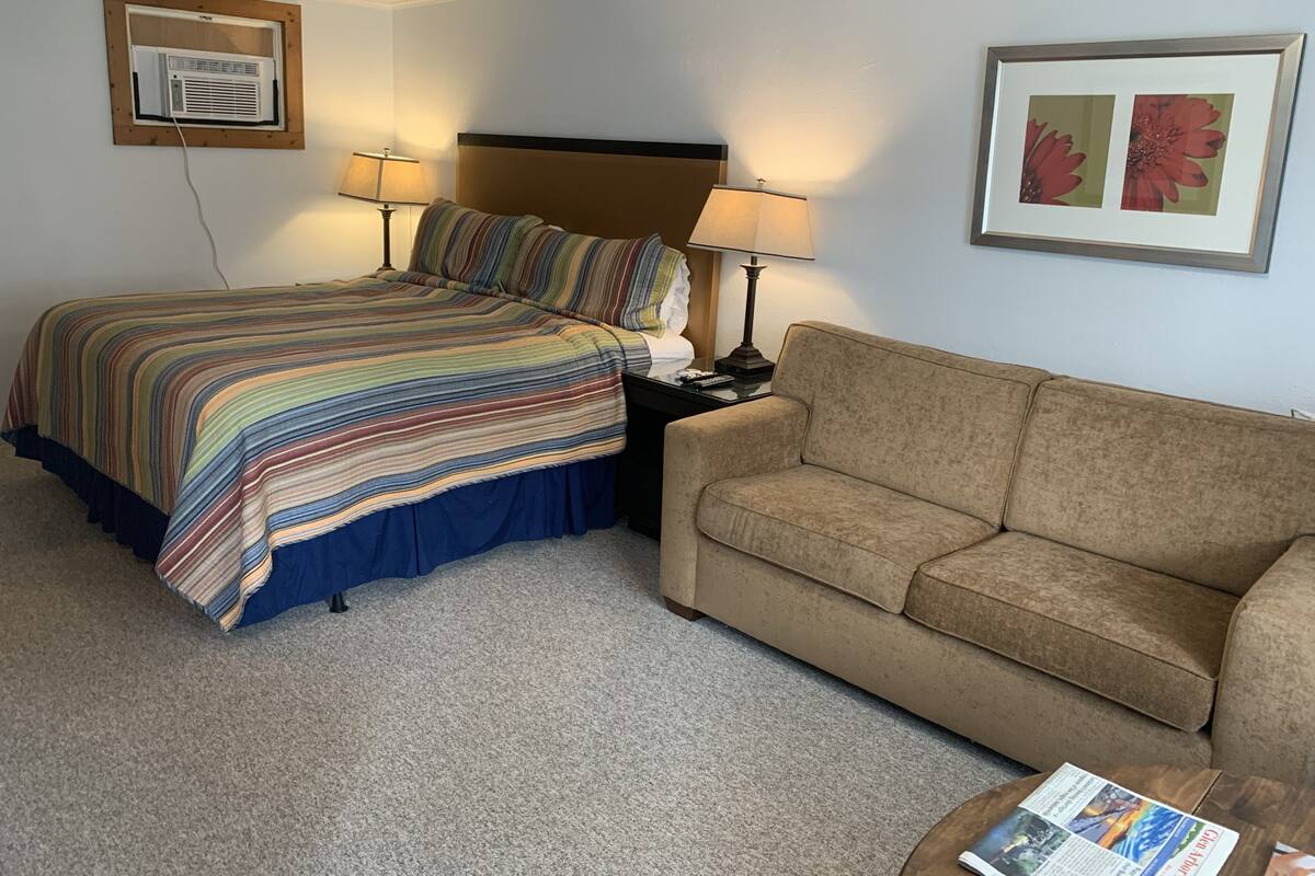 A hotel room with a bed, a couch, two lamps, a framed picture, and an air conditioning unit.