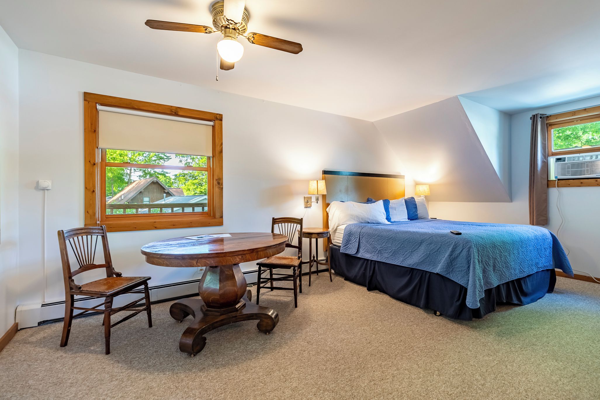 A bedroom with a bed, a table, two chairs, a ceiling fan, and a window with a view of a house.