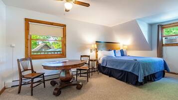 A bedroom with a bed, a table, two chairs, a ceiling fan, and a window with a view of a house.