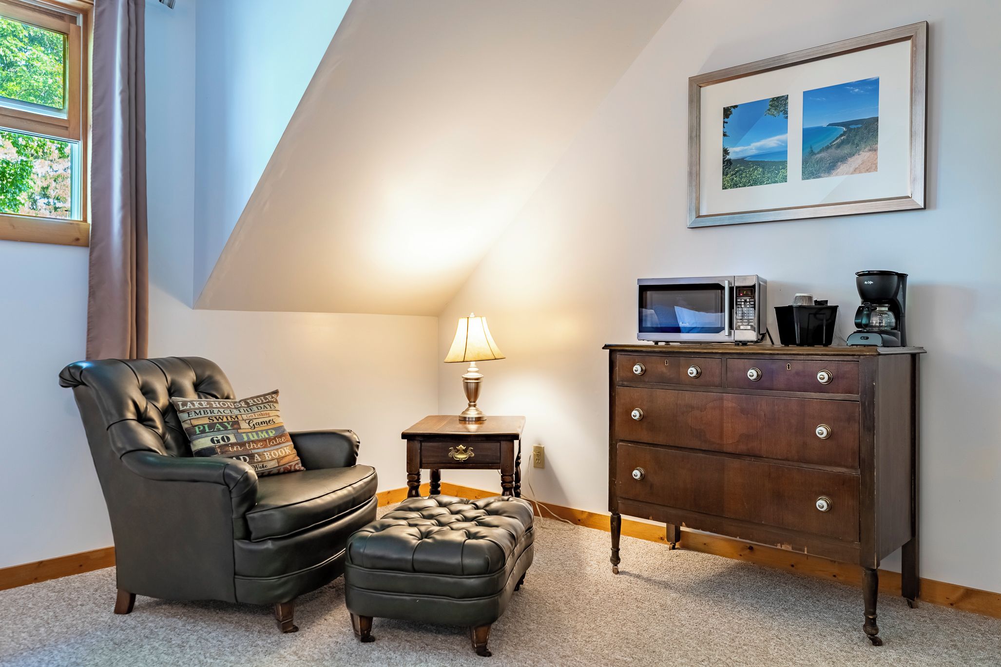 A living room with a leather armchair, ottoman, a small table with a lamp, a wooden dresser with drawers, and a framed picture on the wall.