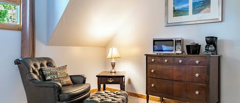 A living room with a leather armchair, ottoman, a small table with a lamp, a wooden dresser with drawers, and a framed picture on the wall.