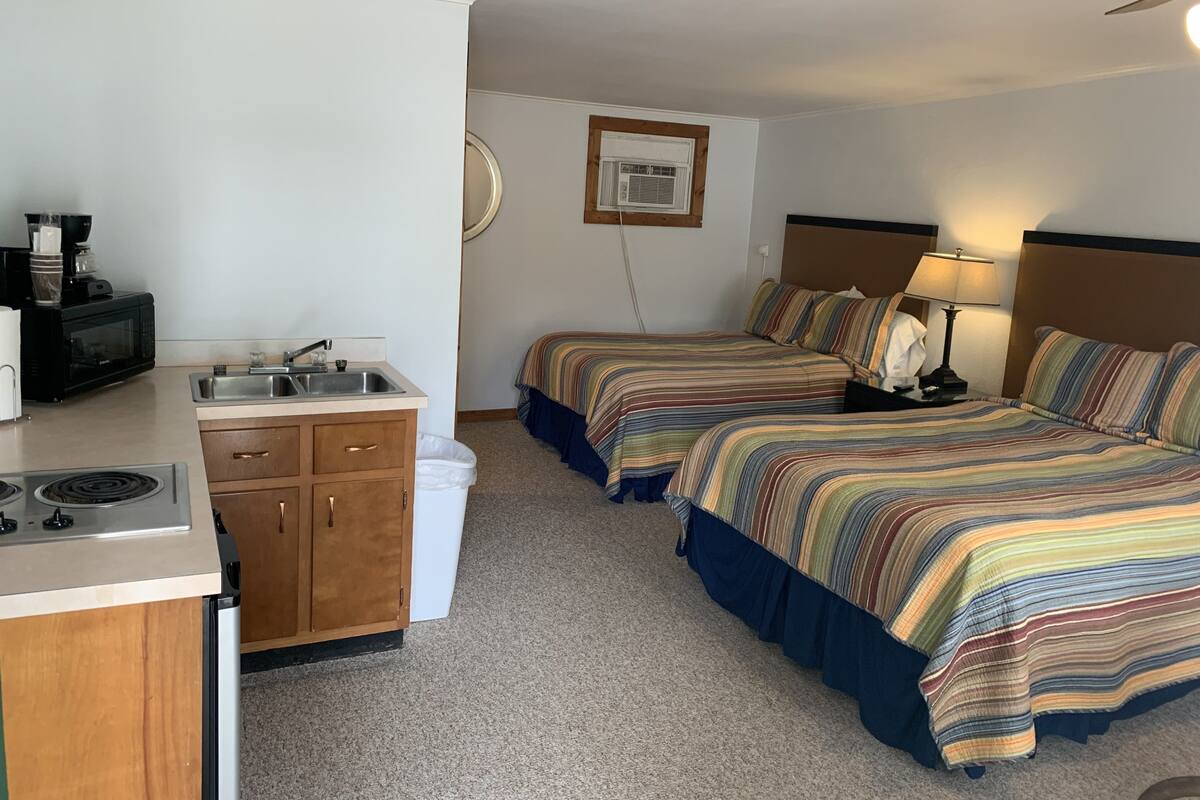 A compact hotel room with a kitchenette, two beds with striped bedding, a ceiling fan, and a wall-mounted air conditioner.