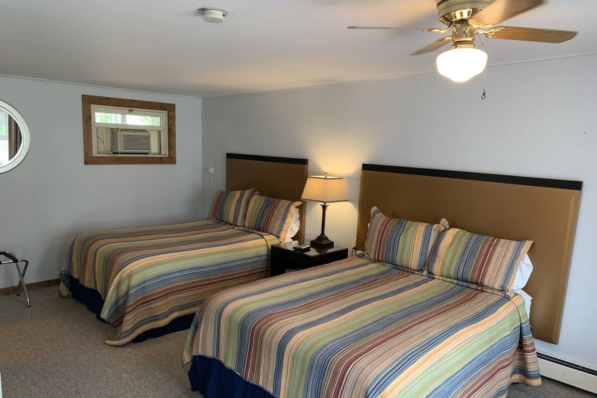 A hotel room with two beds, a ceiling fan, a nightstand with a lamp, a window with blinds, and a wall-mounted air conditioning unit.