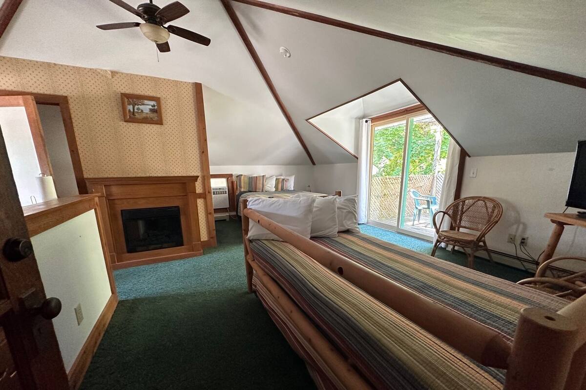 A cozy attic bedroom with a sloped ceiling, a wooden bed, a fireplace, and a large window offering a view of the outdoors.