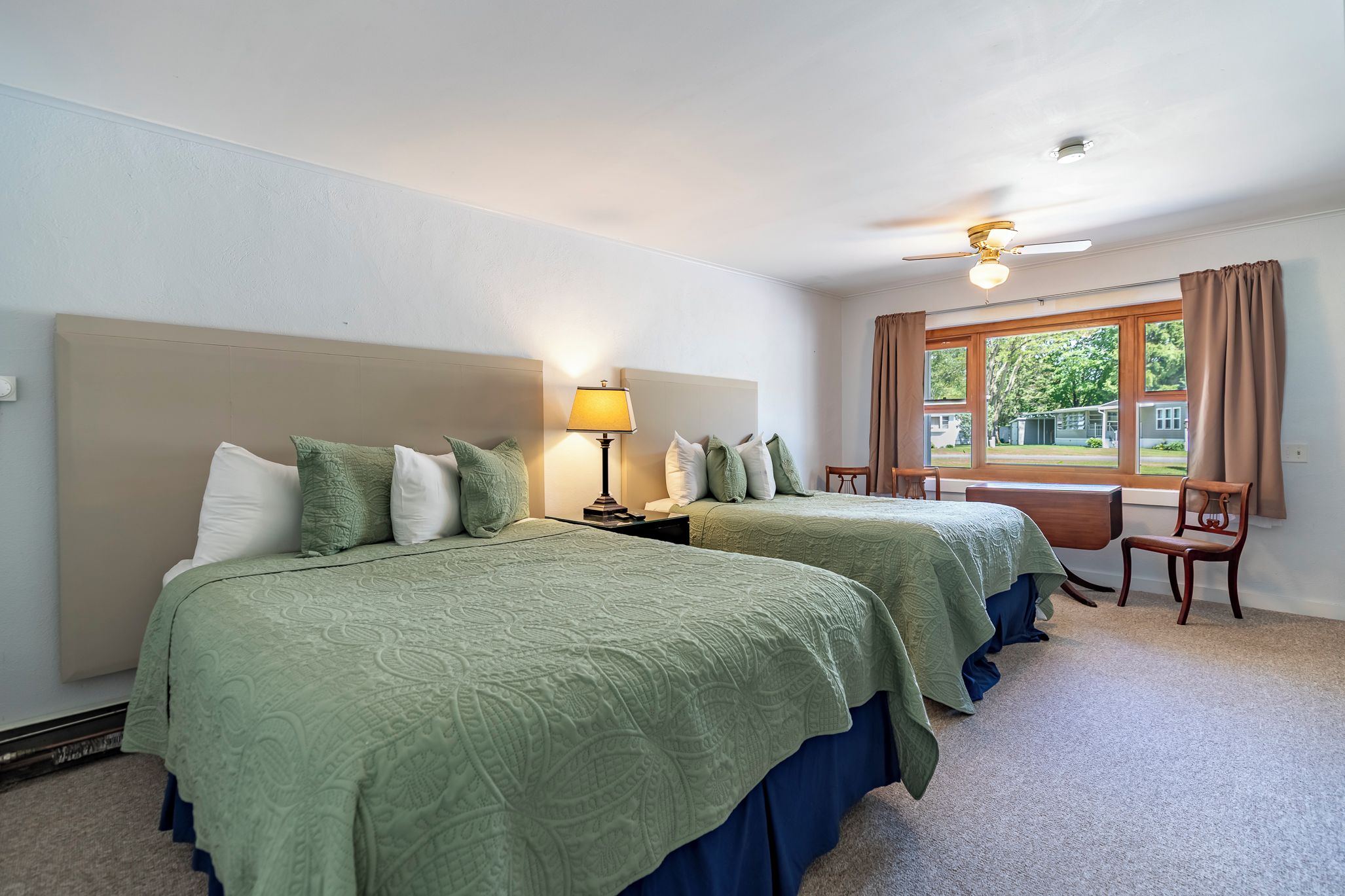 Una habitación de hotel con dos camas, una mesita de noche con lámpara, una ventana con cortinas y un ventilador de techo.