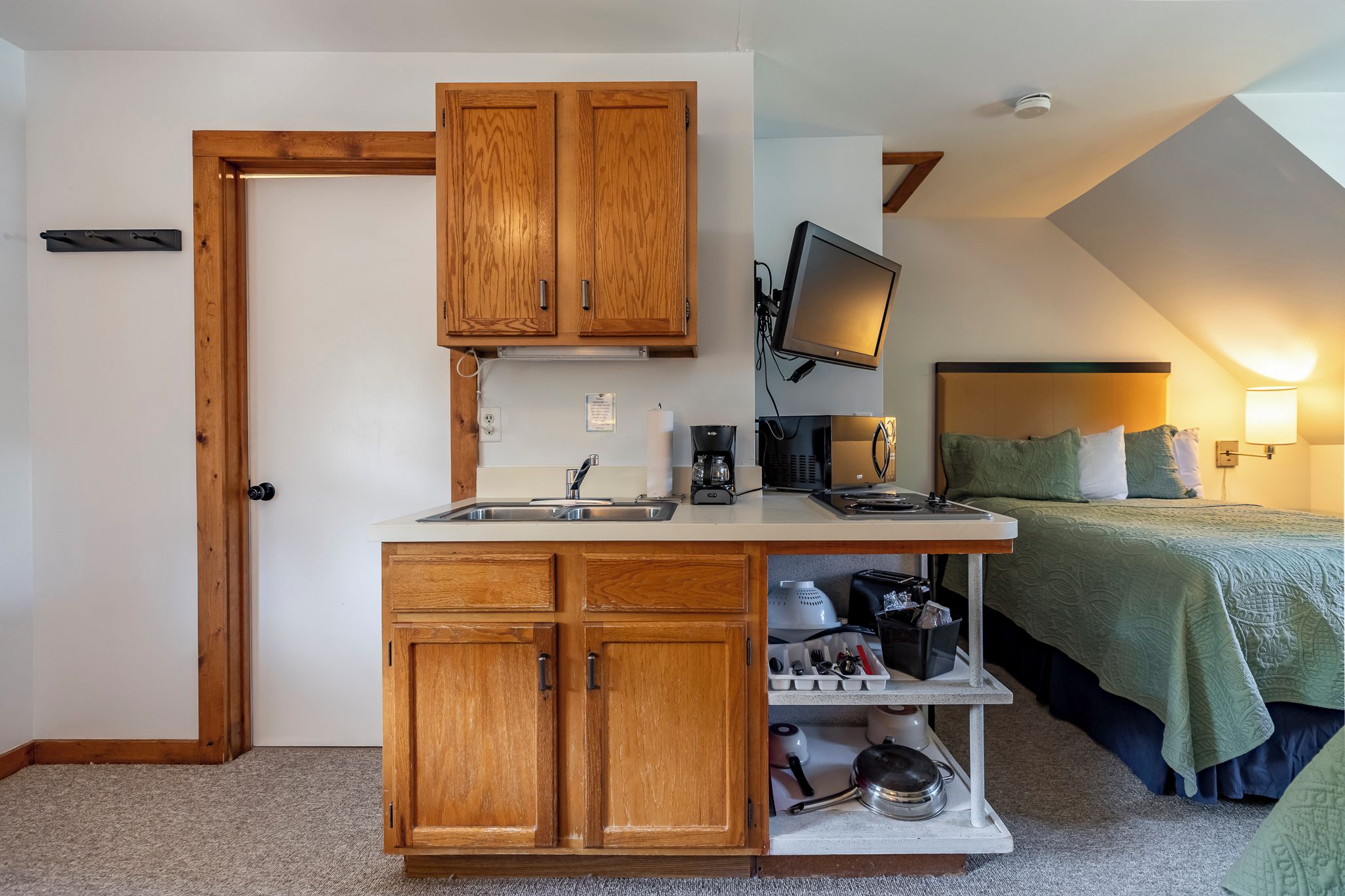 A compact kitchenette with wooden cabinets, a sink, and a countertop. There is a bed with a green blanket and pillows in the adjacent room.