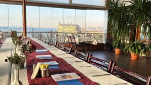 A long table set with a red patterned tablecloth, chairs, and a view of a cityscape through large windows. - Kadikoy Rihtim Hotel (Istanbul)