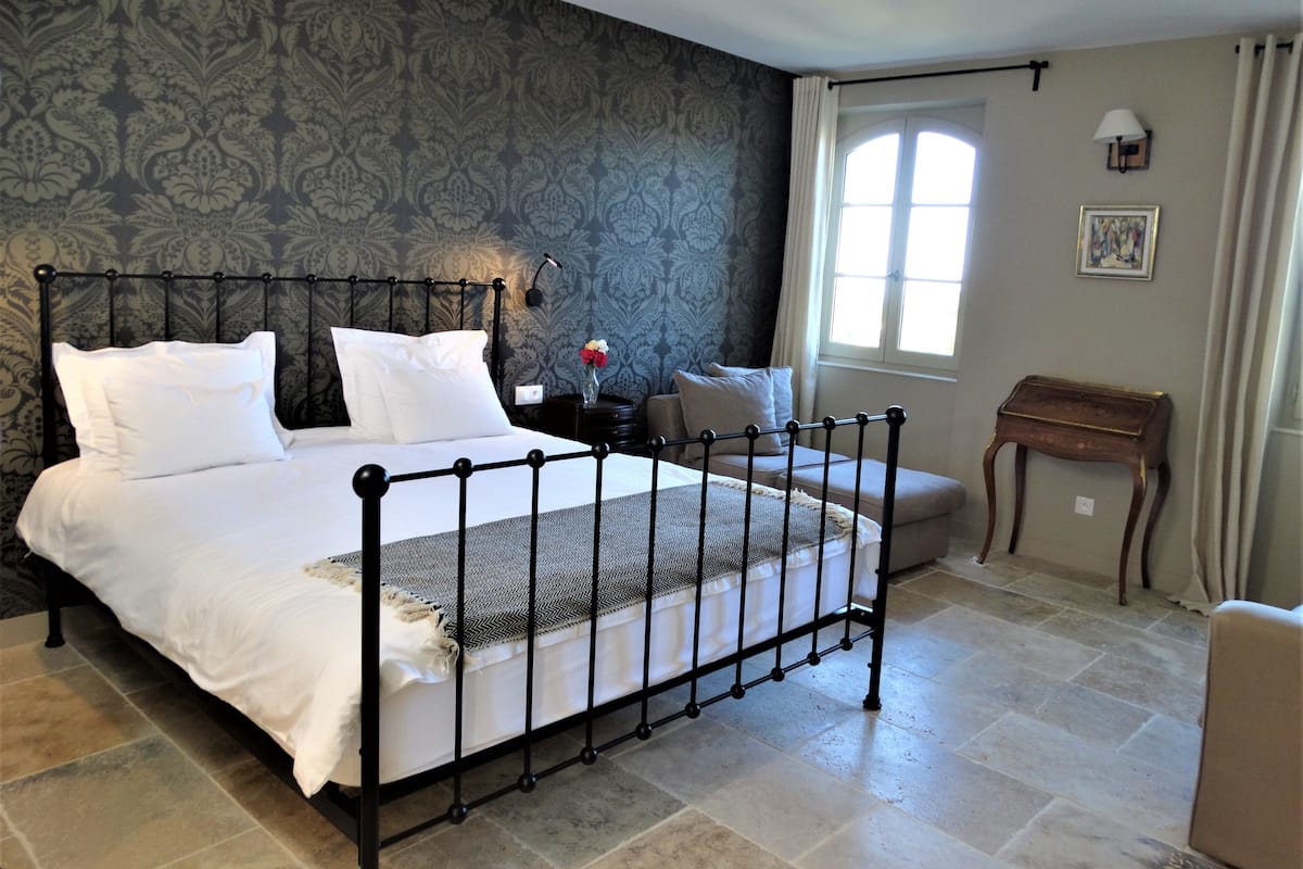 Ein Schlafzimmer mit Bett, Nachttisch, Stuhl, kleinem Tisch, Fenster und Wand mit gemustertem Tapeten.