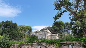 A stone wall with electric vehicle charging stations, a large building with arched windows, and lush greenery in the background. - Chambre d'hôtes Château MontPlaisir (Valreas)