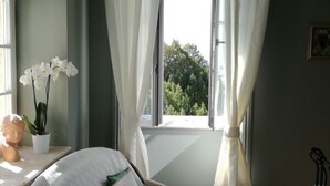 A bedroom with a bed, a window with curtains, a potted plant, and a bust on a shelf. - Chambre d'hôtes Château MontPlaisir (Valreas)