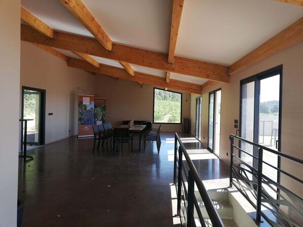 A spacious room with a high ceiling and exposed wooden beams, a dining area with tables and chairs, and large windows offering views of the outdoors. - Chambre d'hôtes Château MontPlaisir (Valreas)