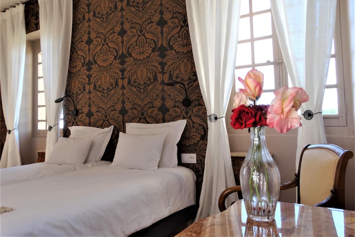 Ein Hotelzimmer mit einem Bett, weißen Vorhängen, einem gemusterten Wandpapier und einer Vase mit Blumen auf einem Tisch.