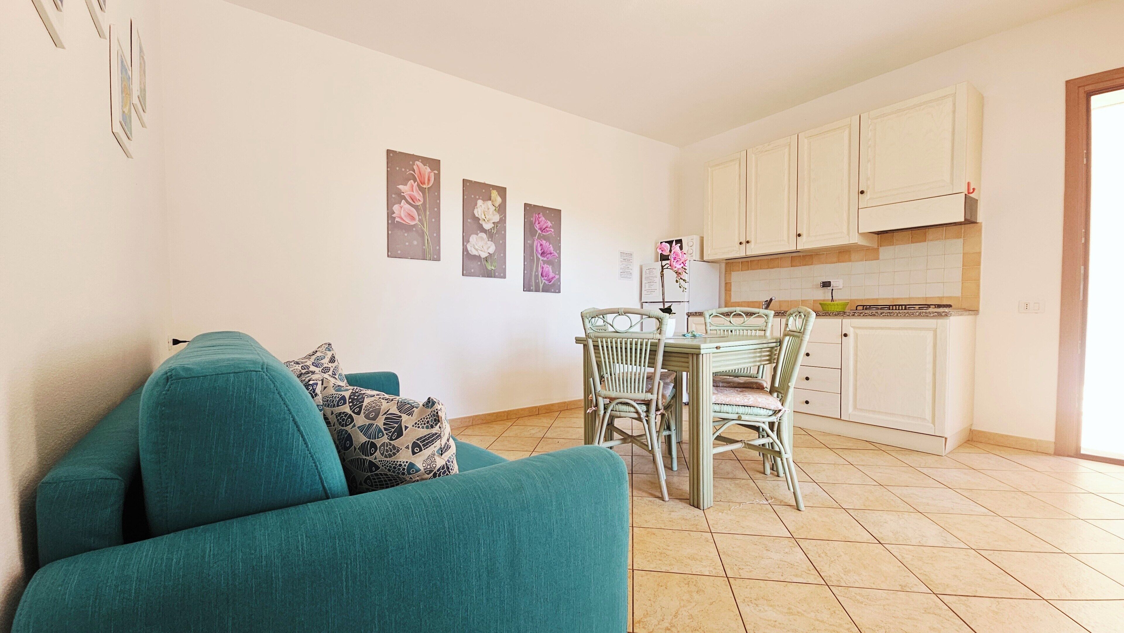 Una sala de estar con un sofá azul aguamarina, una mesa de comedor con sillas y una zona de cocina con armarios.