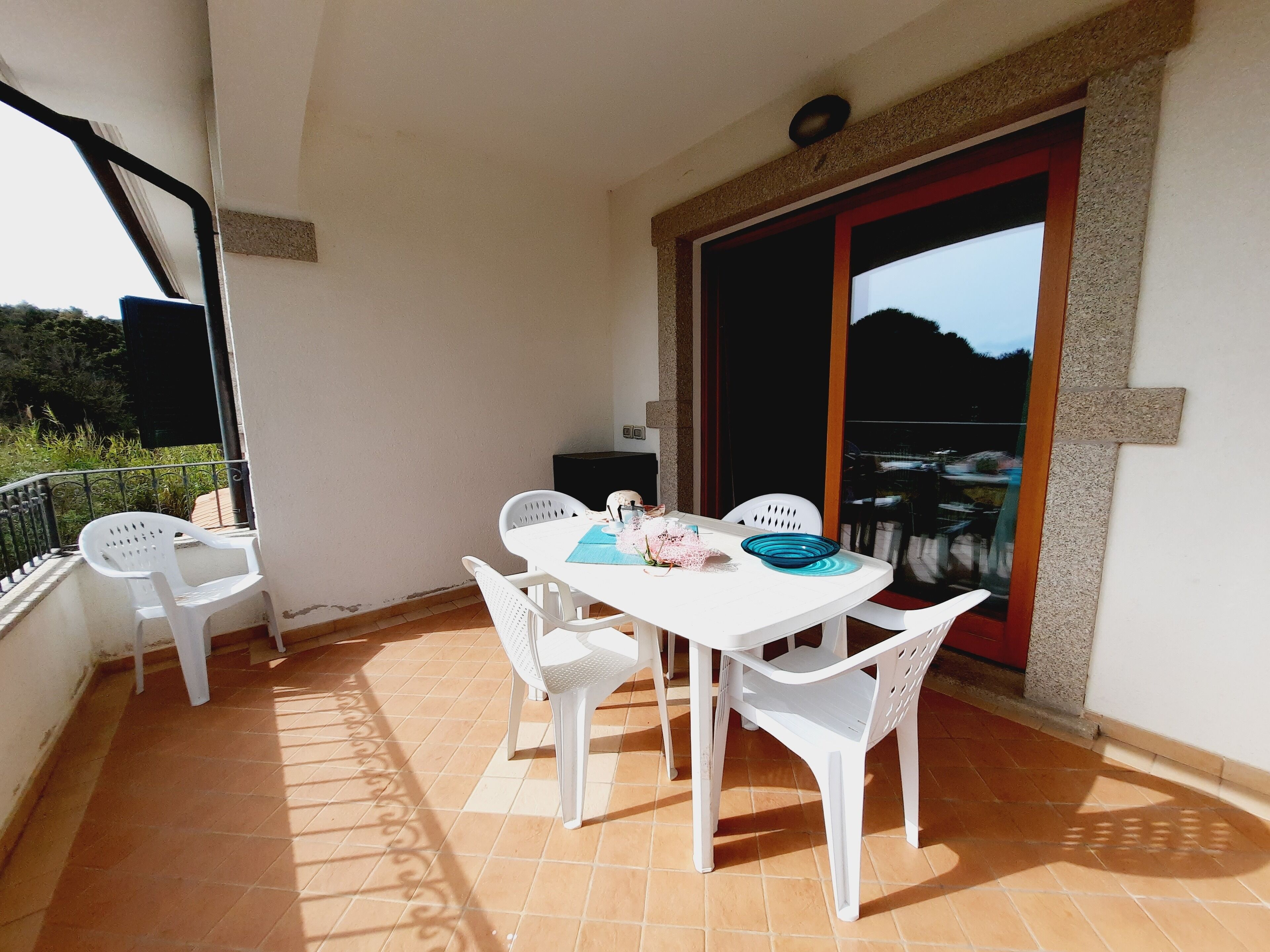 Un balcón con una mesa y sillas blancas, una puerta con marco rojo y vistas al exterior.