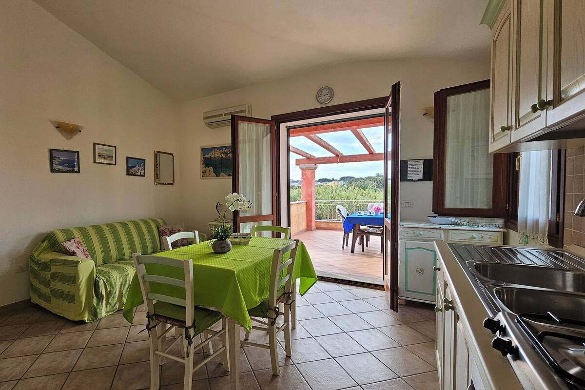 A cozy kitchen with a dining area, a green-striped sofa, a table with a green tablecloth, and a view of the outdoors through an open door.