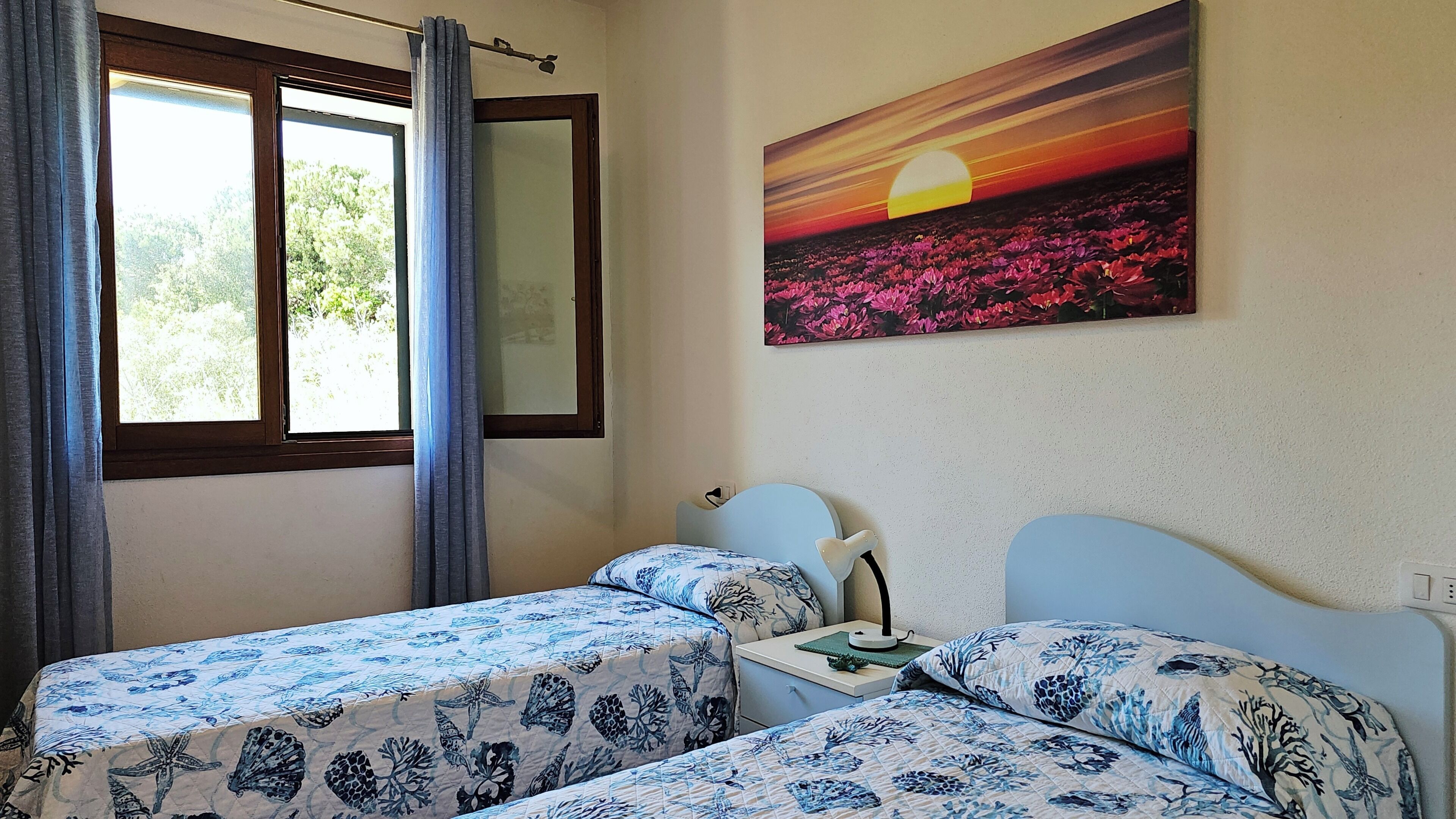 Une chambre avec deux lits, une table de chevet, une lampe, une fenêtre avec des rideaux et un tableau représentant un coucher de soleil accroché au mur.