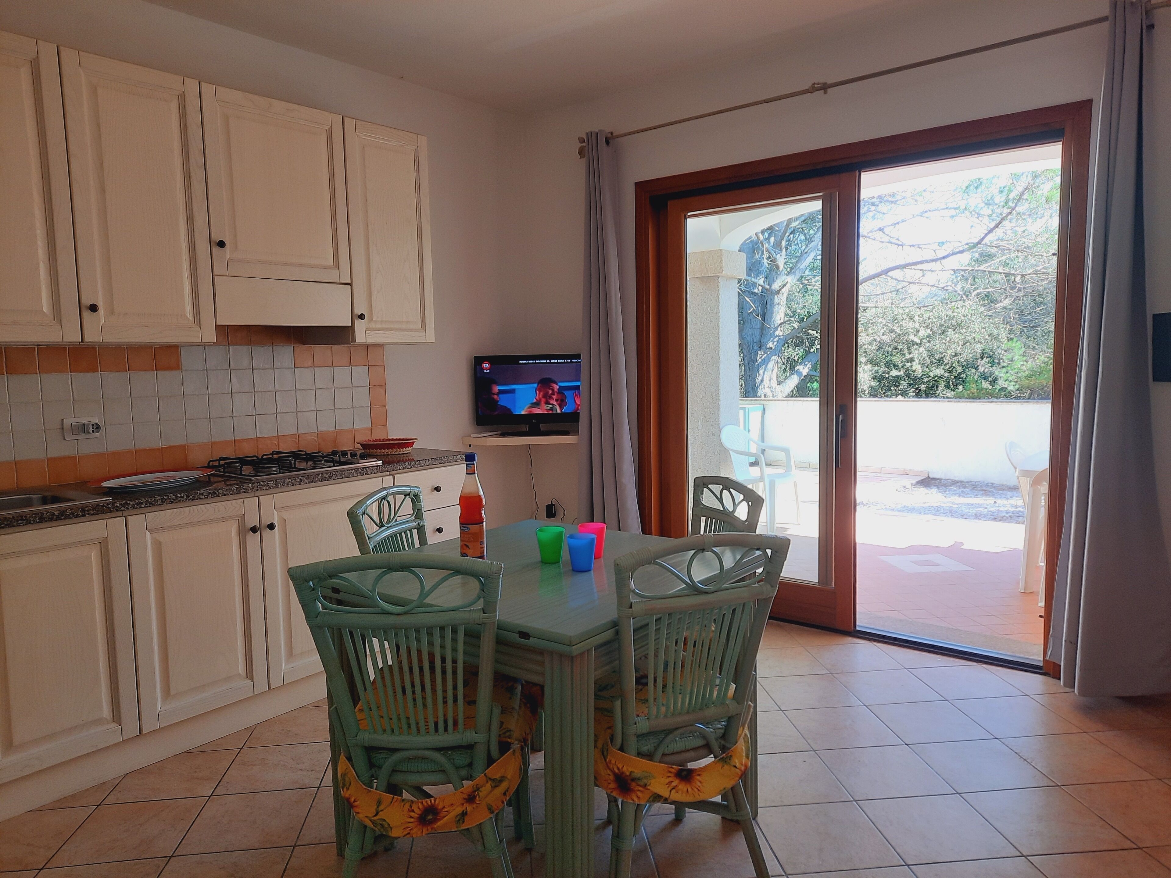 A kitchen with white cabinets, a gas stove, and a television on a shelf. There are two green chairs with floral seat covers and a table with a few items on it.