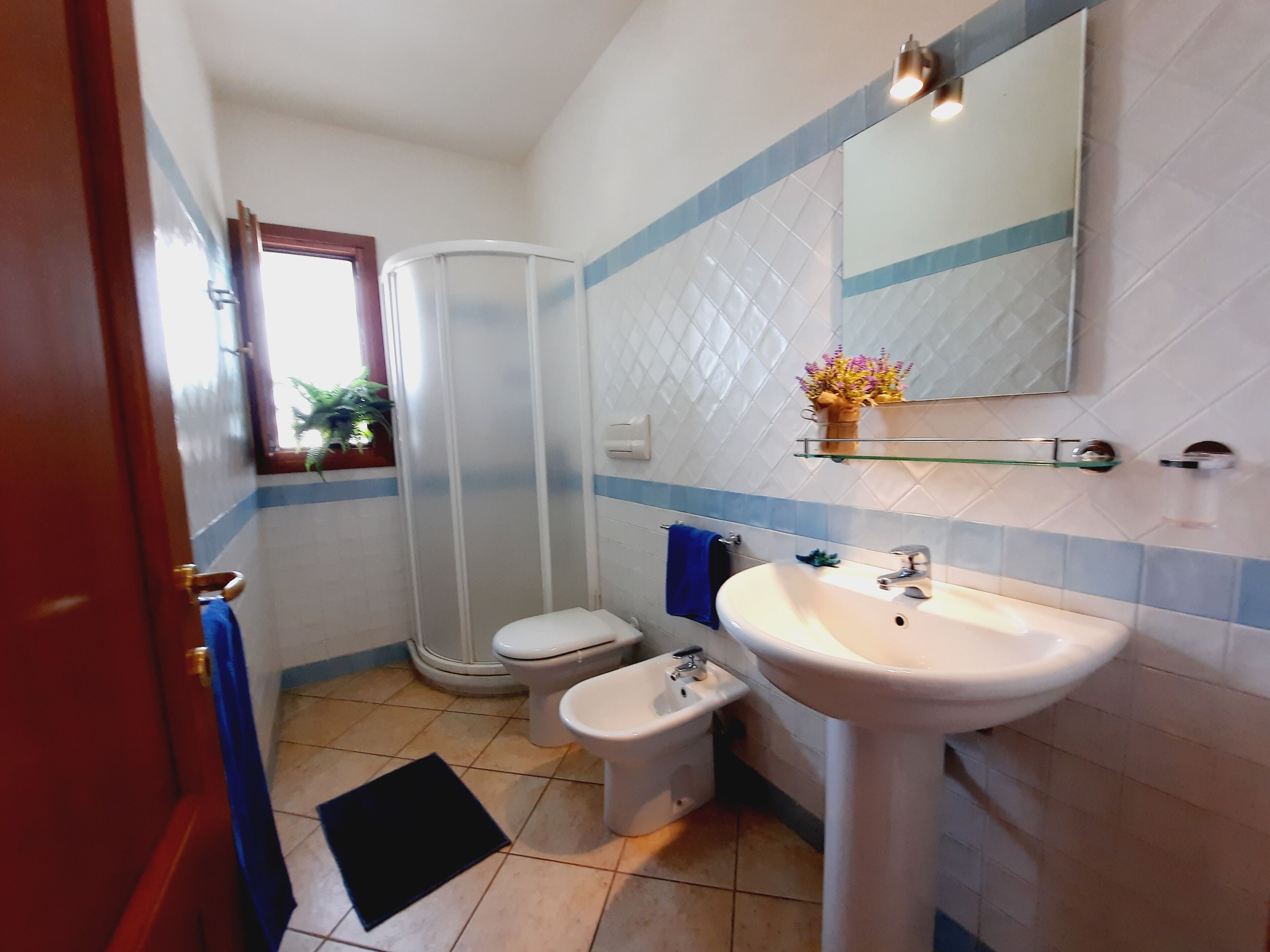 Ein Badezimmer mit Waschbecken, Toilette, Bidet und Dusche. Über dem Waschbecken befindet sich ein Spiegel und ein Fenster mit Pflanzen davor.